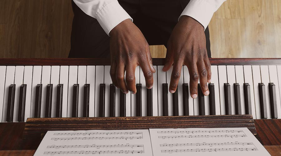 The hands and wrists of a Black pianist wearing a white cuffed shirt are posed on top of piano keys with sheet music