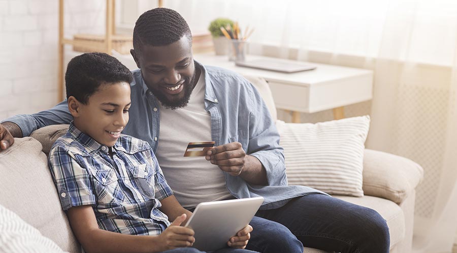 An African American man sits on a couch with a credit card, beside an African American boy using a tablet