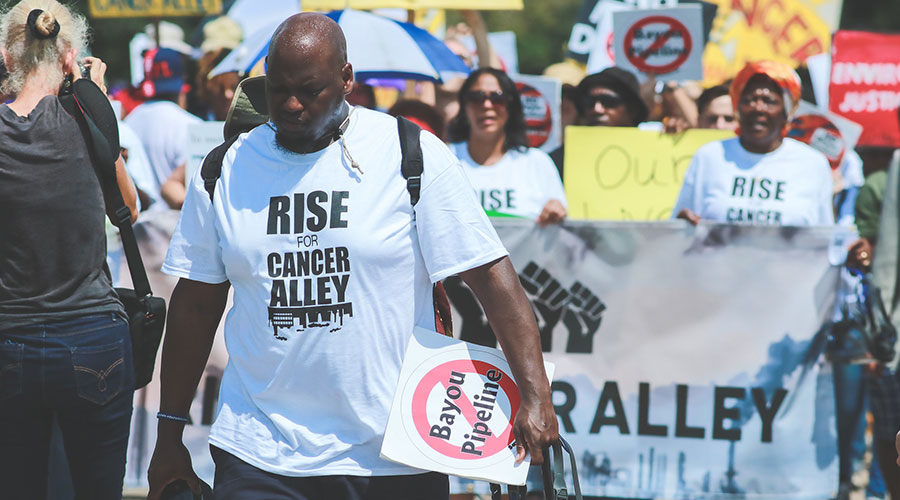 A Black man wears a t-shirt that reads Rise Up for Cancer Alley while walking in a protest march.