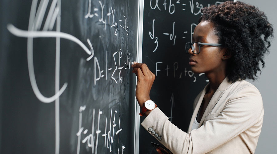 An African American woman stands at a chalkboard, solving a complex math problem.