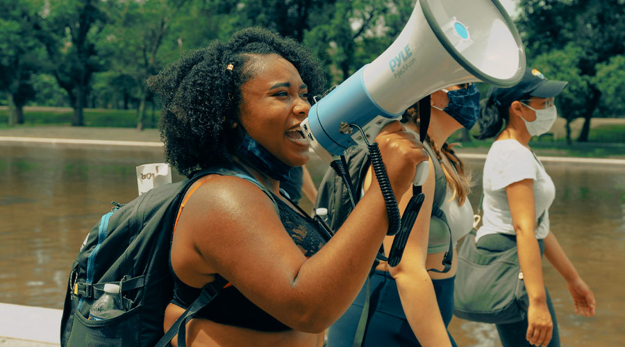 A young African American woman speaks into a megaphone while walking with two other women wearing face masks.