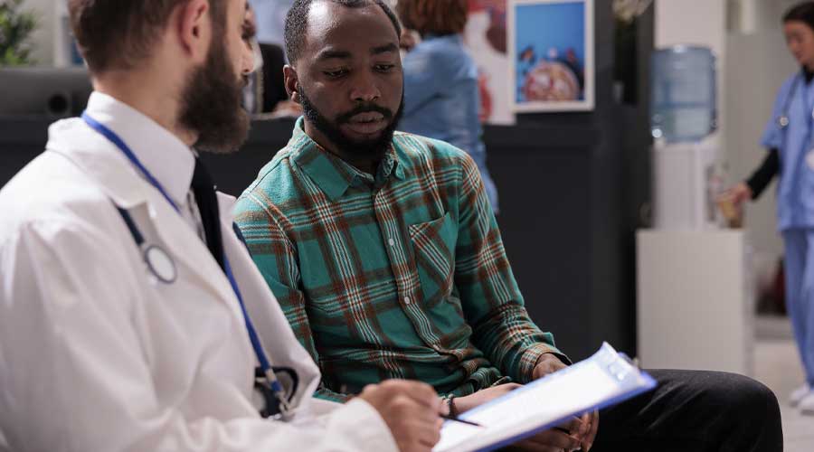A Black man speaking with a doctor in a waiting room that is holding a clipboard.