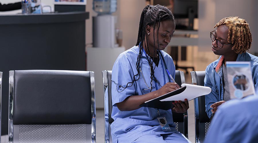 A female African American medical professional wearing blue scrubs sits with another African American woman and fills out a form in a hospital setting.