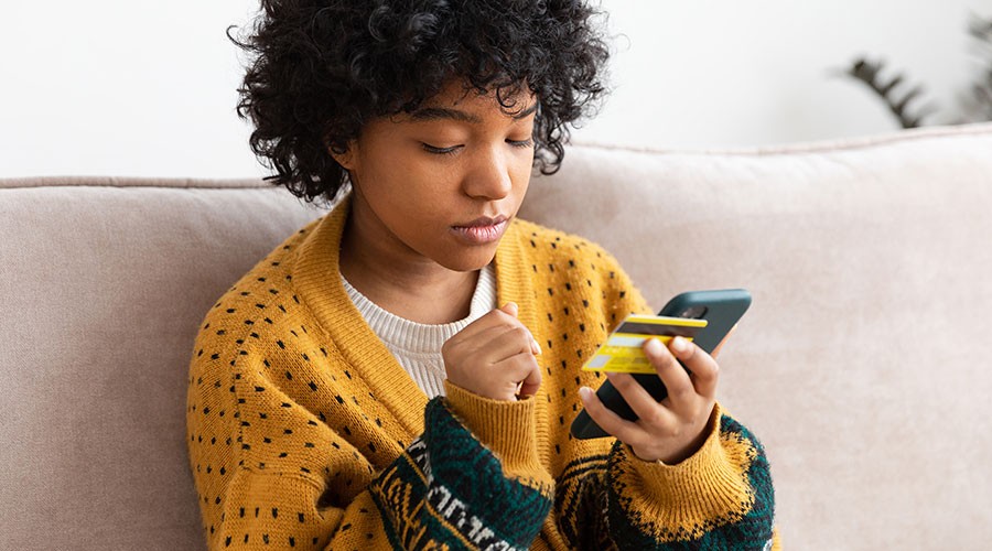 Image of a Black American sitting on a couch looking at a cell phone and holding a yellow credit card.