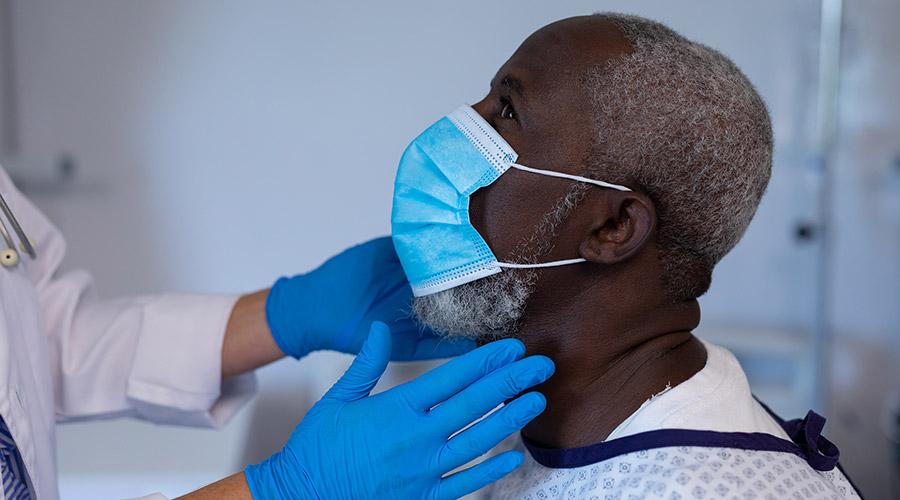 A Black man with a mask on tilts his head up as a doctor examines his neck