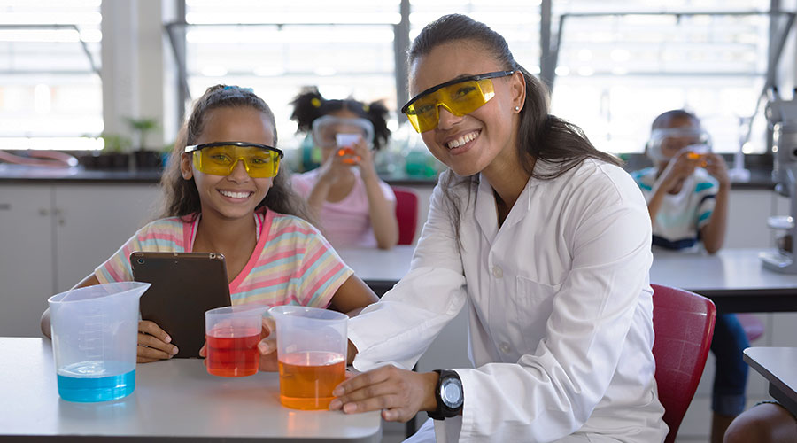 A teacher wearing a white lab coat sits next to a young girl as they perform a science experiment