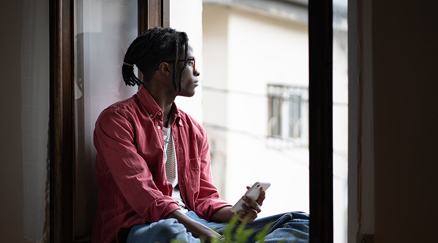 Image of a Black individual in a striped shirt and red button down holding a phone and sitting while looking out a window