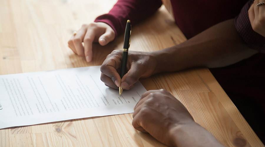 Image of a Black person signing a document with a pen on a wooden table next to another person