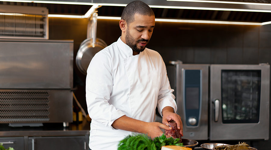 A Black man wearing a chef’s coat in the kitchen while he prepares food