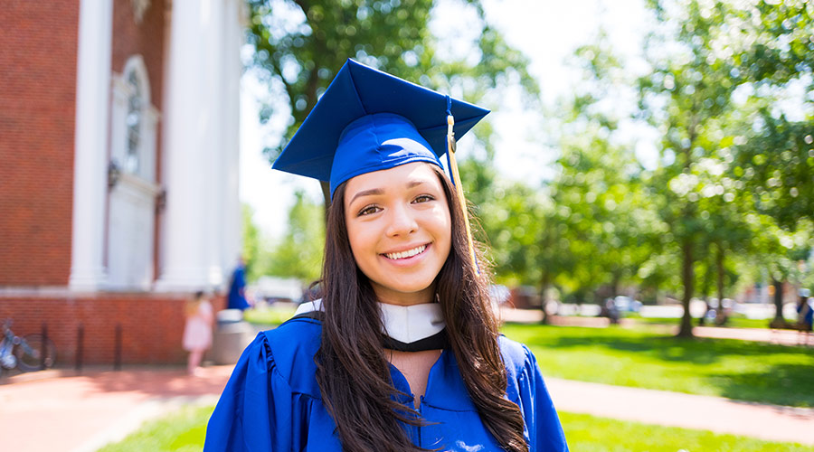 Female college graduate standing and smiling while looking into the camera wearing a blue cap and gown