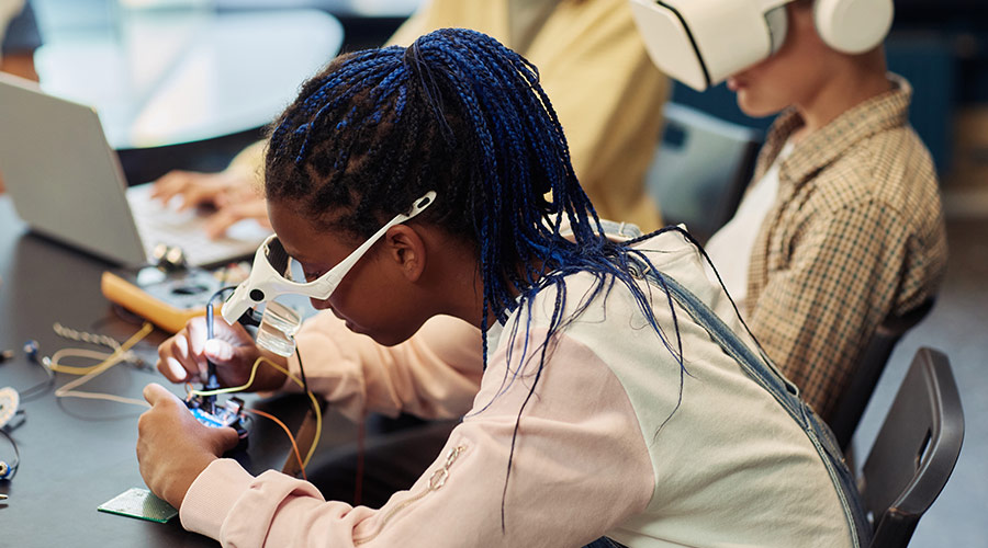 A Black woman in STEM wearing goggles and inspecting a device in a STEM classroom.