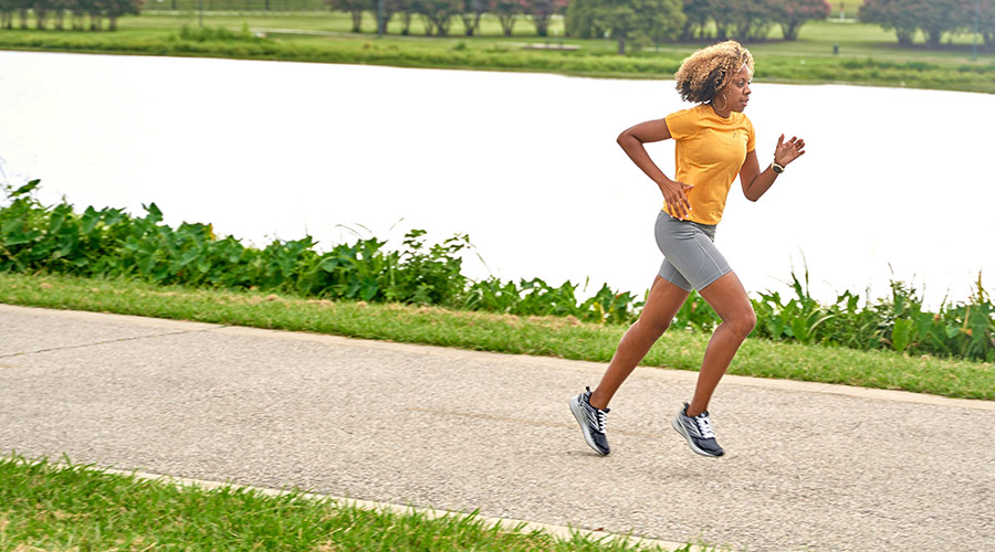 Black woman, wearing a yellow t-shirt and gray shorts, running on a trail near a lake