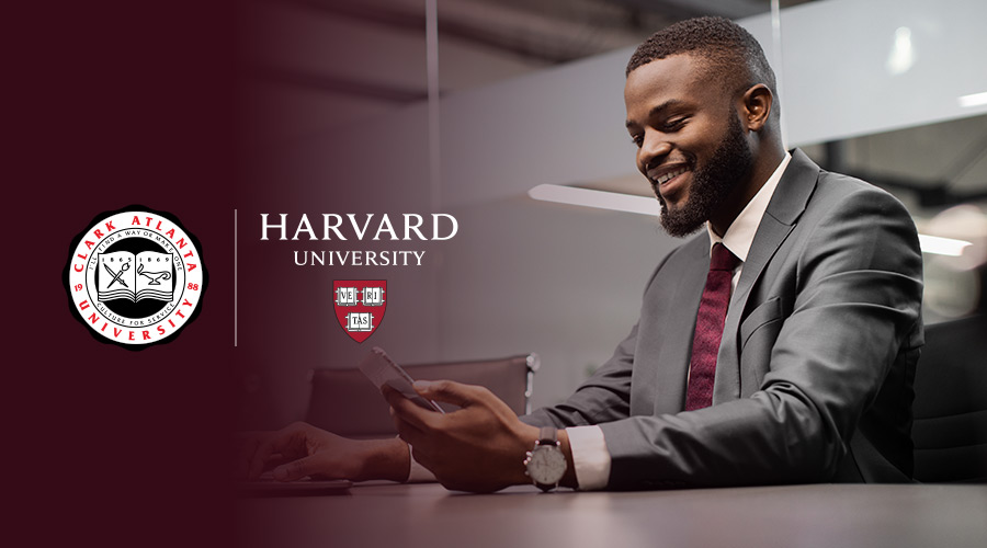 A Black man sitting down, dressed in a suit and holding his phone with the Harvard University and Clark Atlanta University logos appearing to the left of the man