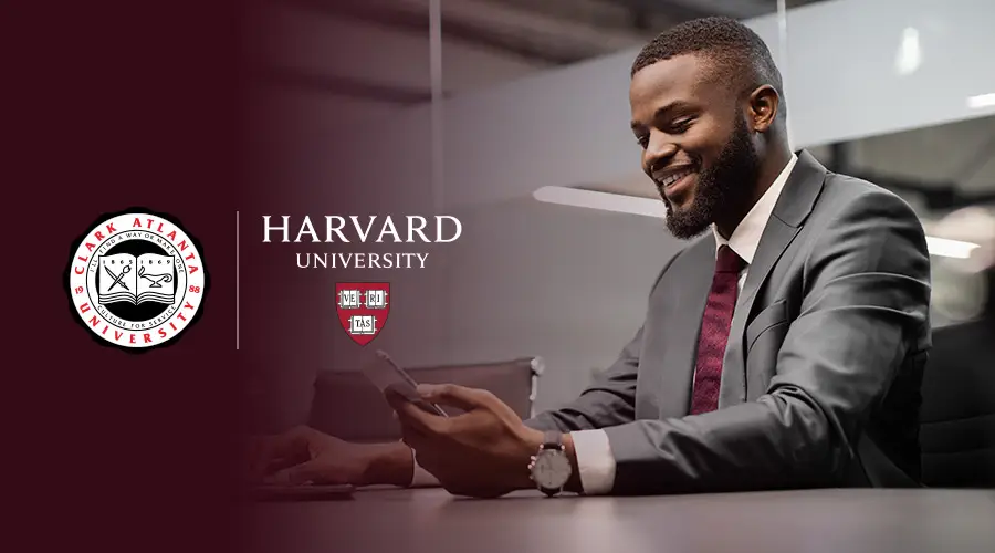 A Black man sitting down, dressed in a suit and holding his phone with the Harvard University and Clark Atlanta University logos appearing to the left of the man