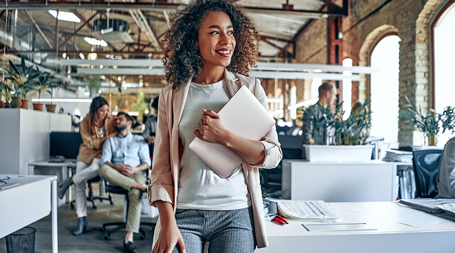 A Black woman in business attire holding a laptop in an office