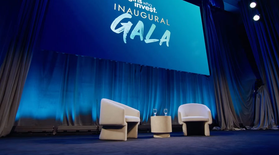 A stage with blue curtains, two white chairs and a table at the 2024 Girls Who Invest Inaugural Gala