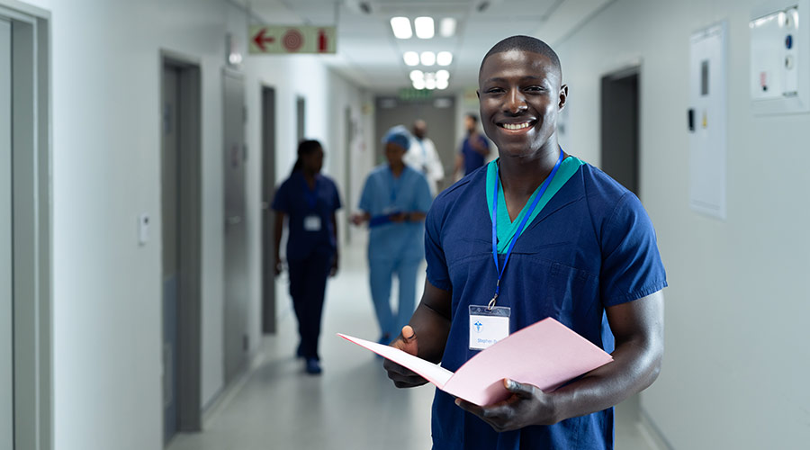 Black male nurse smiling while holding a folder and standing in a hospital hallway