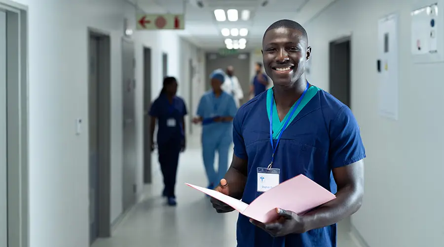 Black male nurse smiling while holding a folder and standing in a hospital hallway