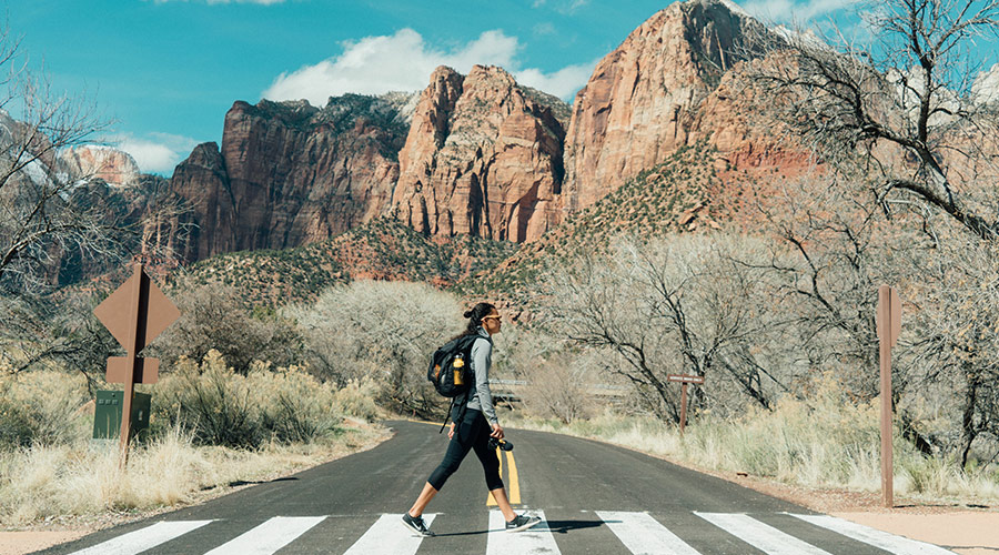 A hiker wearing leisure wear and a backpack walking in a crosswalk at a national park with rocky formations in the background