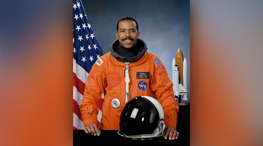 Dr. Bernard Harris in his orange NASA space suit, standing in front of the American flag.