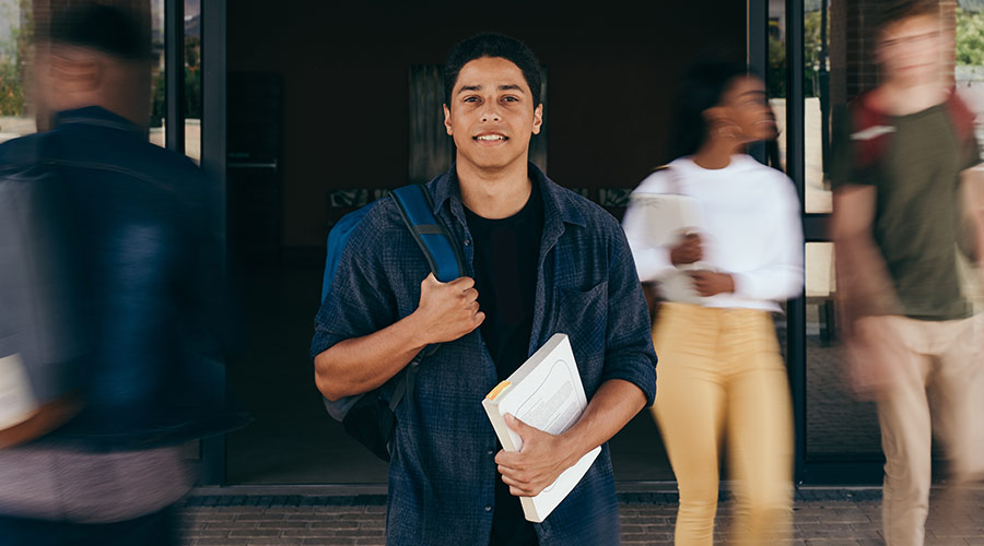 Young student in a black t-shirt and dark blue button down holding a backpack and notebook