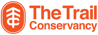 Orange oval with a white tree-shaped symbol in the middle next to the text "The Trail Conservancy" in orange.