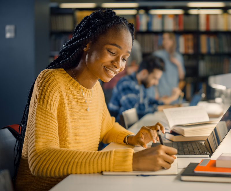 Young woman in a yellow sweater sitting at a desk using a laptop and writing in a notebook.