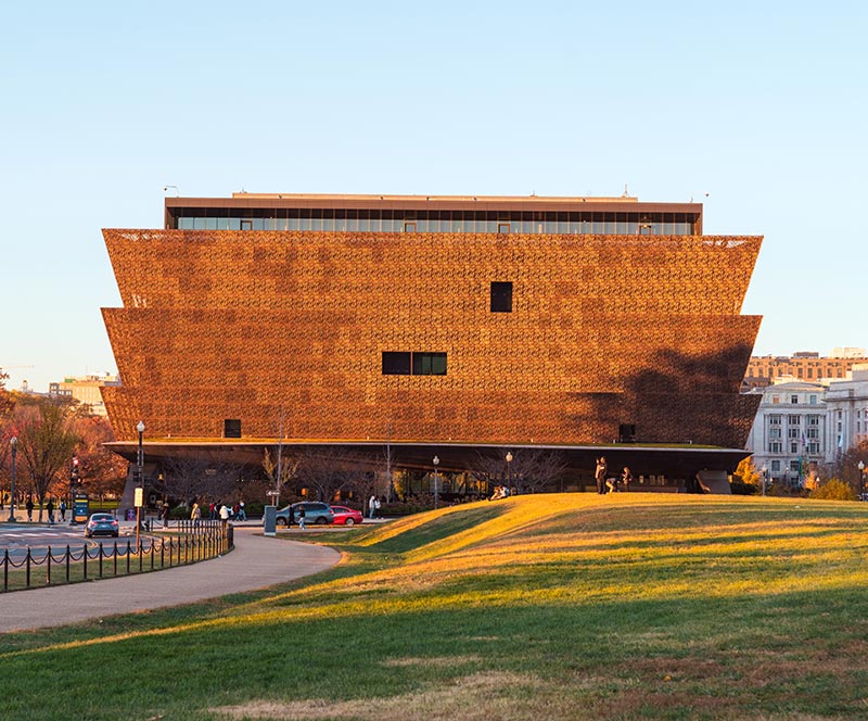 The facade of the National Museum of African American History and Culture (NMAAHC) with a blue sky in the background and a grassy hill and sidewalk in the foreground.