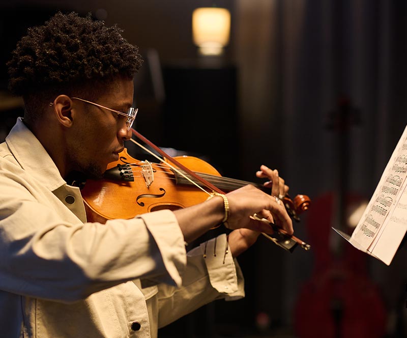 Young man wearing glasses and a tan button-down shirt playing a violin in front of a sheet music stand.