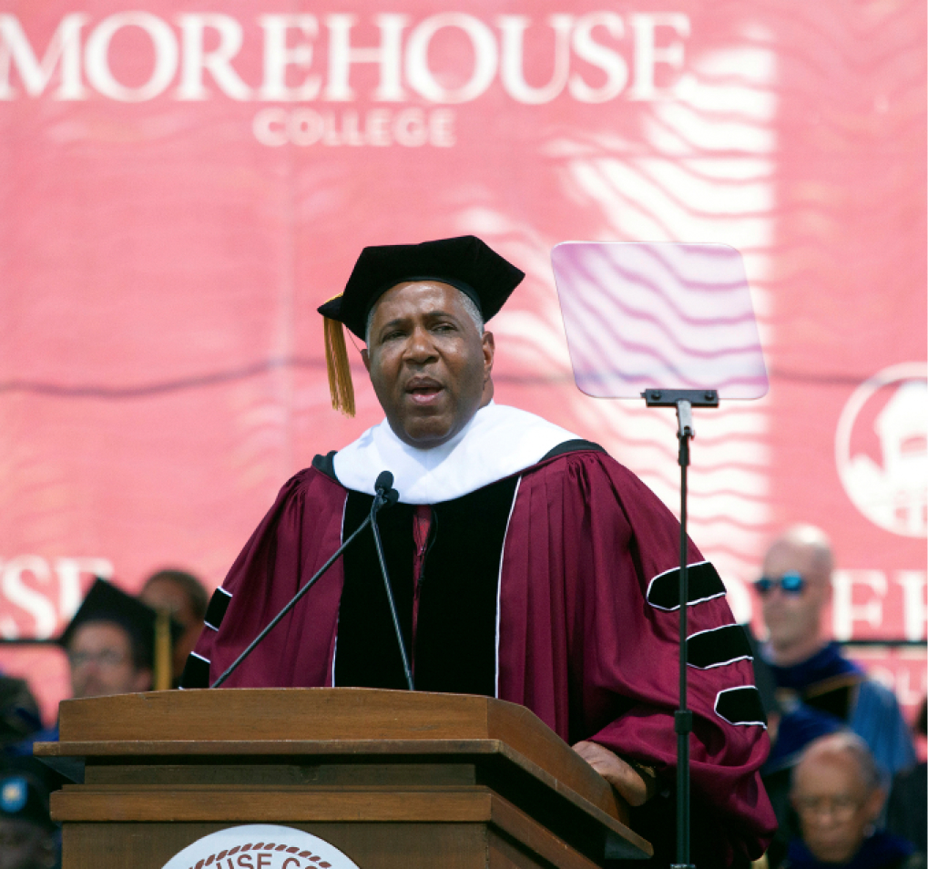 Robert F. Smith, wearing a maroon robe, black cap and stole, stands at a podium speaking into a microphone, with a red Morehouse College banner in the background.
