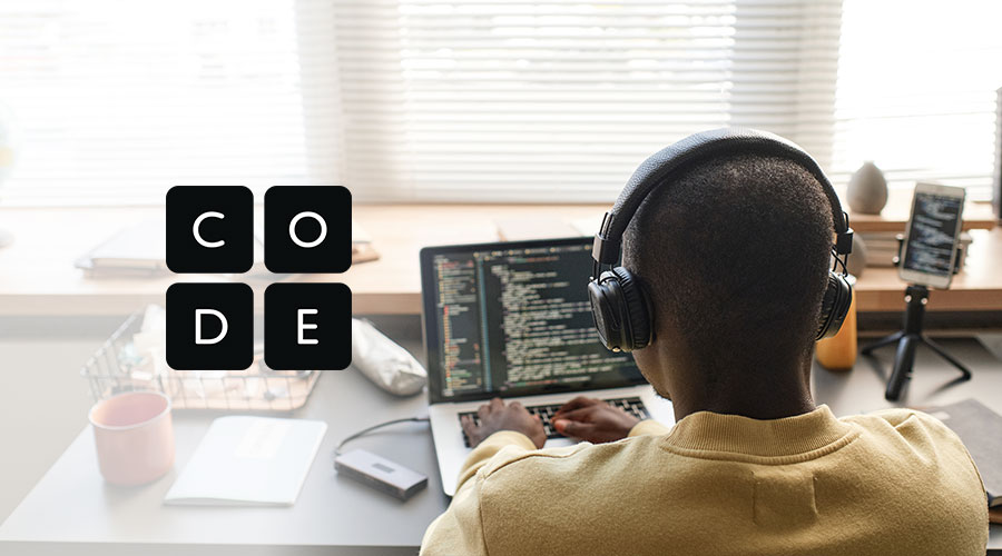 A young man in a yellow sweater, wearing black headphones, sits at a desk coding on his laptop, with the Code.org logo displayed on the side of the image.