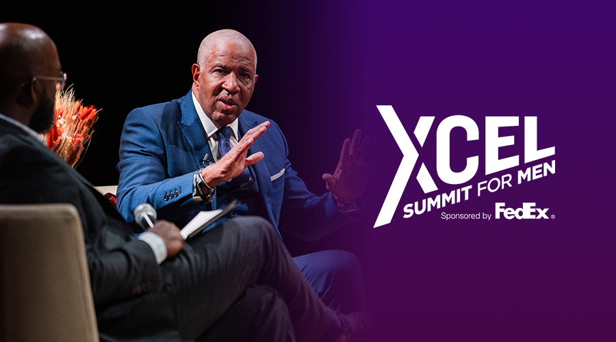 Smith speaks to a host on a stage next to a logo for the XCEL Summit For Men on a purple background.