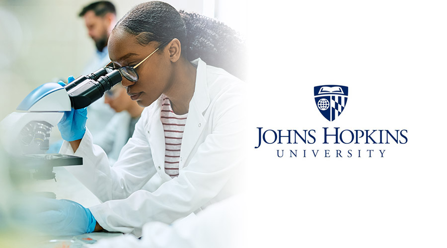 A female student in a white lab coat looking into a microscope next to the logo of Johns Hopkins University.