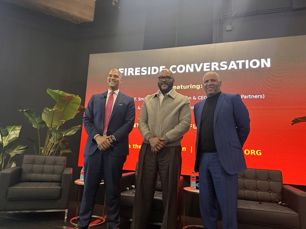 Frank Holland in a blue suit, Tyler Perry in a tan jacket and Robert F. Smith in a blue suit on stage with a red background.