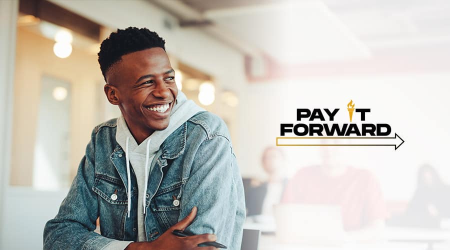 A young man sitting in a hoodie and jean jacket with his arms crossed next to a “Pay It Forward” logo.