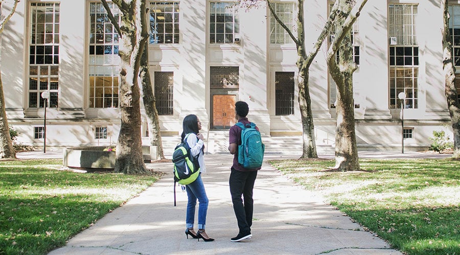 Two individuals wearing backpacks and standing in front of a building on a path facing the entrance.