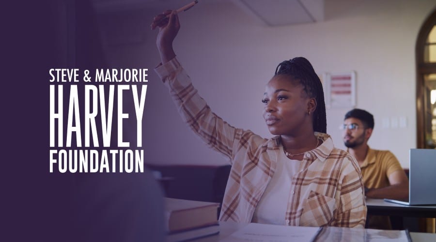Student raising hand in a classroom featuring the Steve & Marjorie Harvey Foundation logo